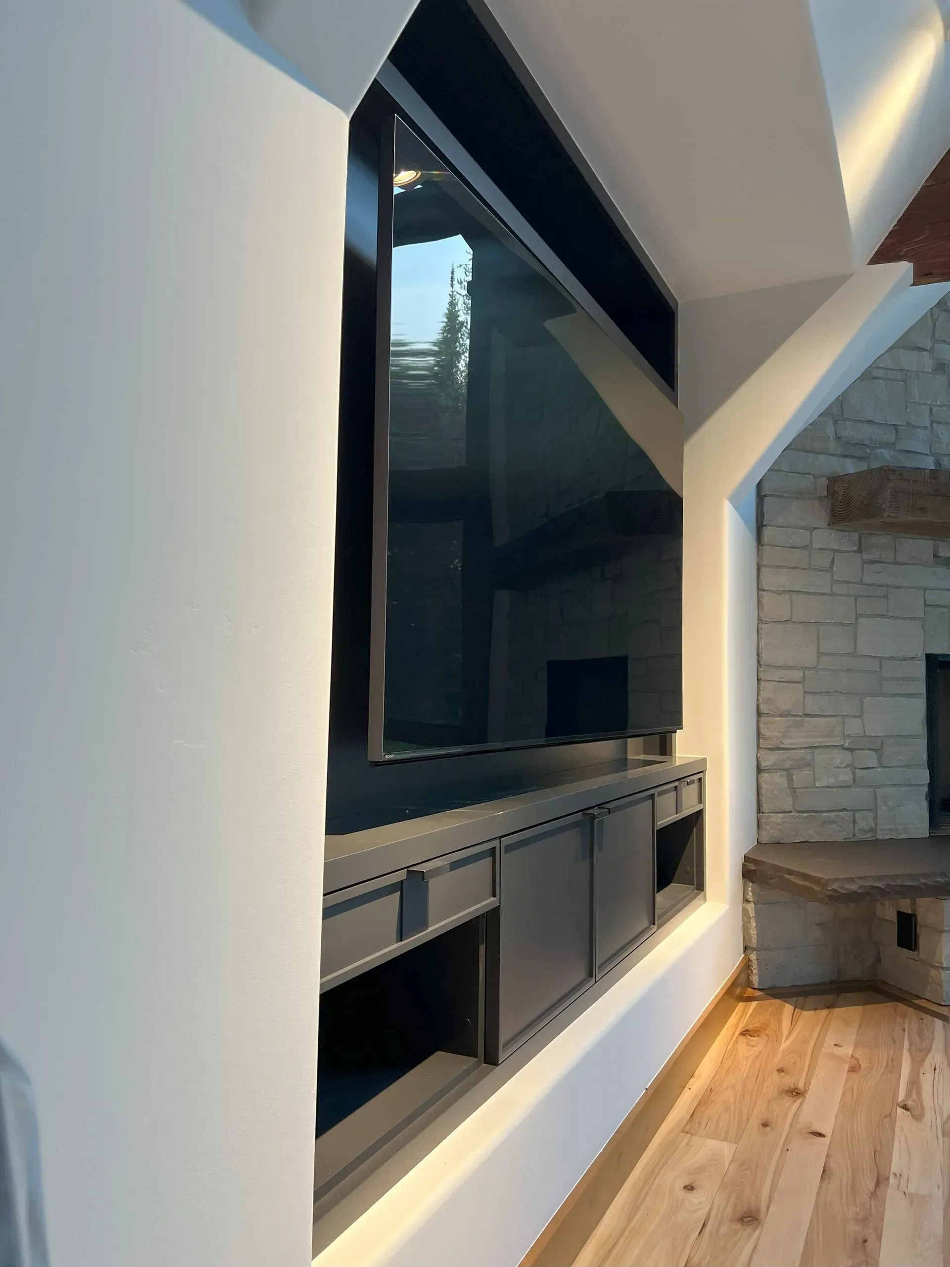 A large, dark flat-screen TV mounted in a recessed wall, set above custom dark cabinetry next to a stone fireplace.