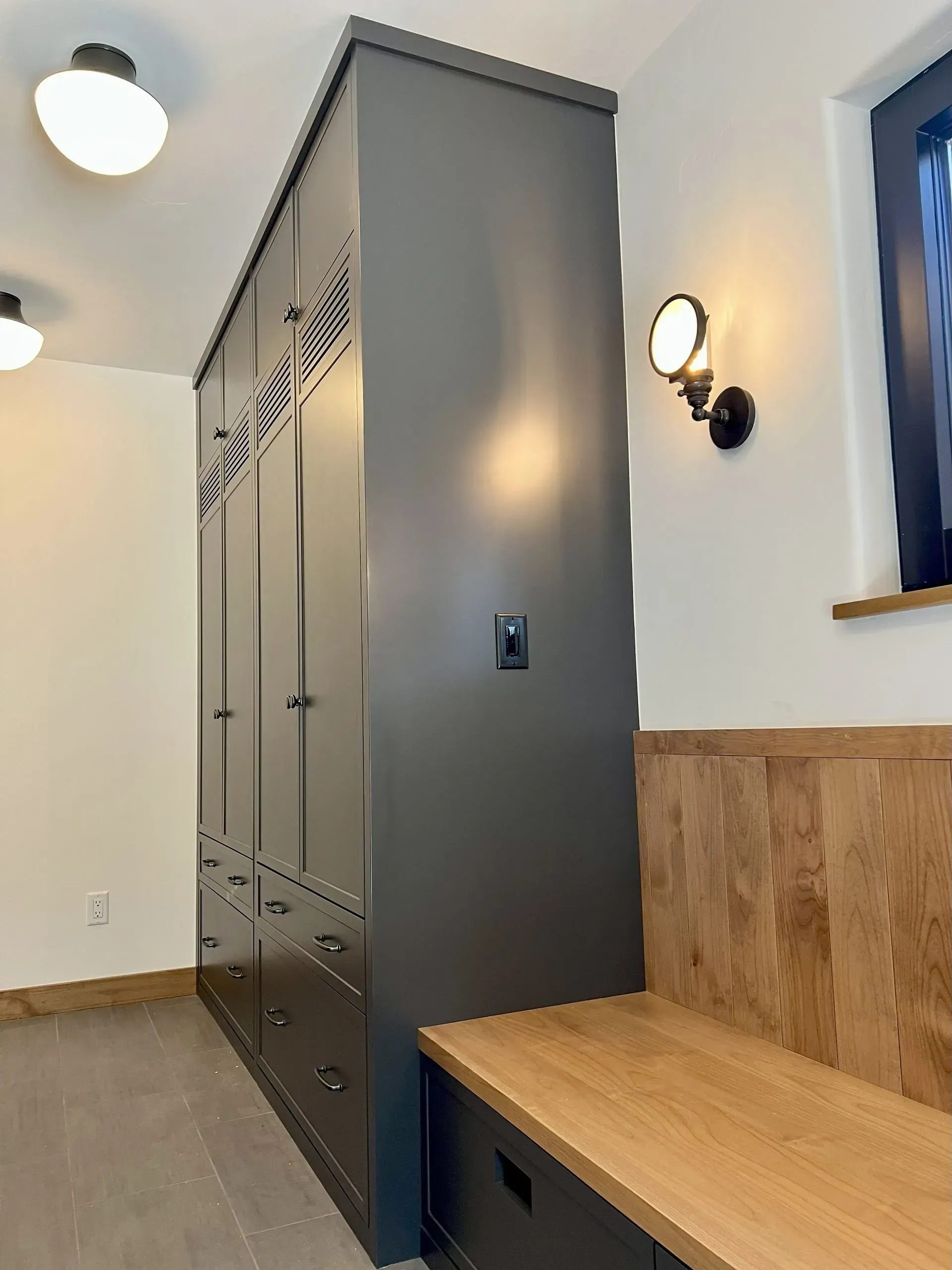 A built-in dark gray locker unit next to a matching wooden bench with drawers in a bright room with wall sconces.