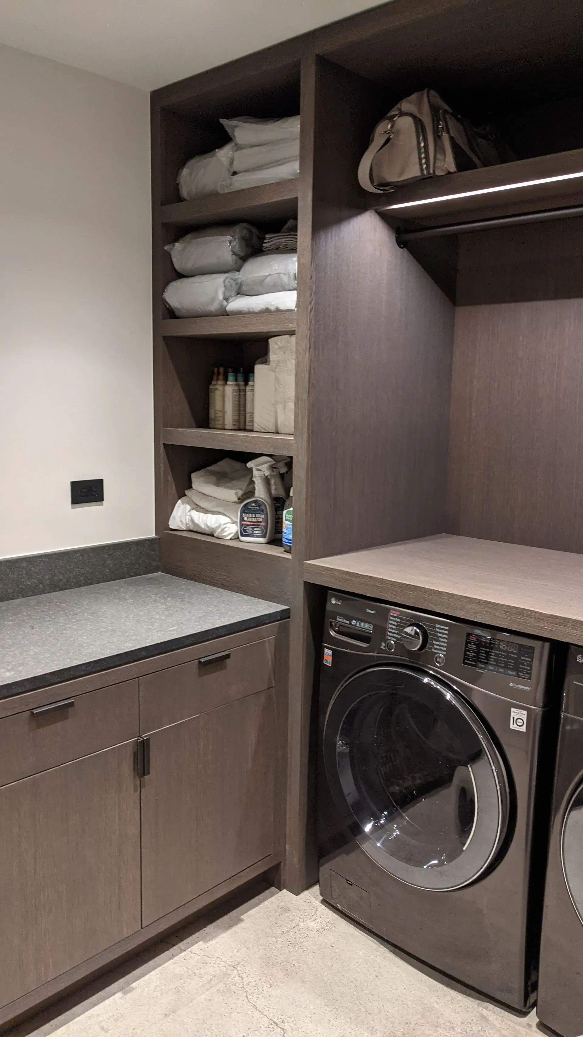 A laundry room with dark wood cabinetry, a grey stone countertop, stacked open shelving with linens, and a dark dryer.