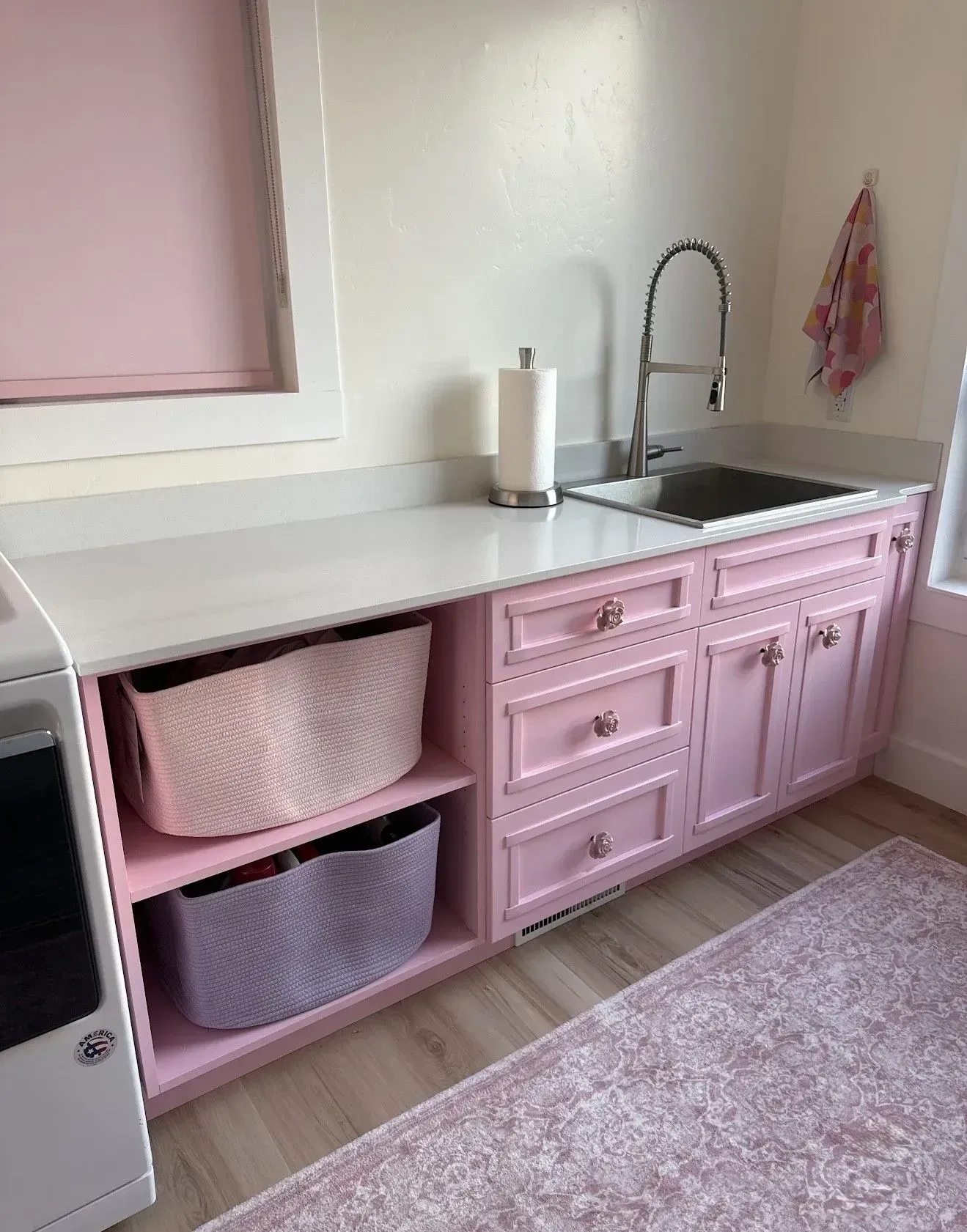 A pink laundry room vanity with a white countertop, sink, open shelving with two woven baskets, and pink cabinetry.