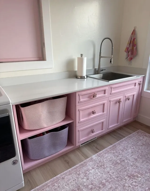 A pink laundry room counter with a white top, open shelving with two baskets, a sink, and matching pink cabinets.