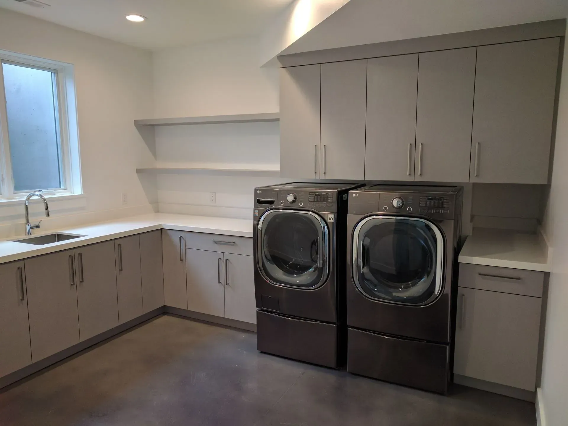 Modern laundry room with gray cabinets, a sink, open shelving, and two dark gray front-loading machines.