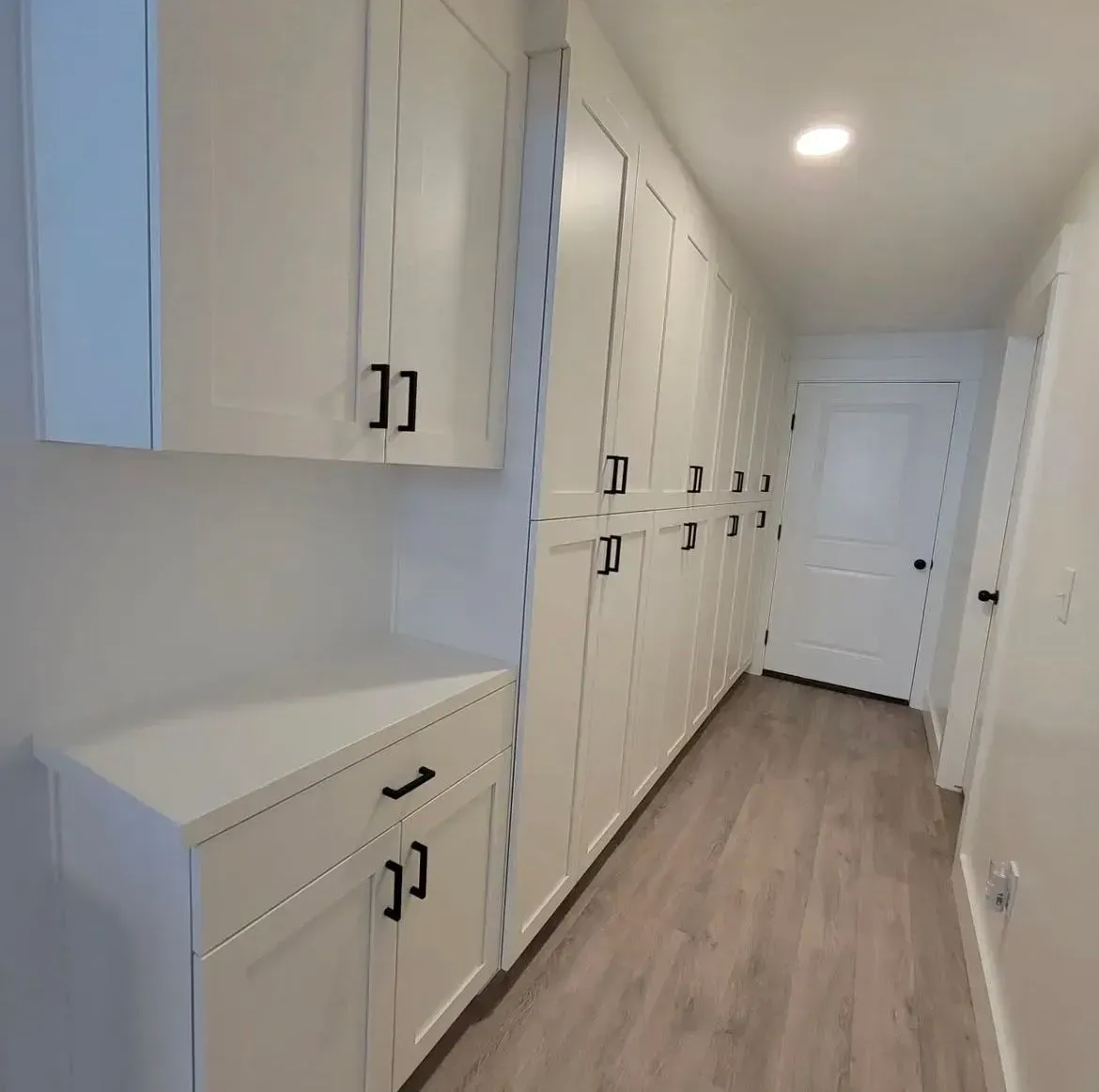 A hallway with white cabinetry along one wall, a white countertop, and light wood-look flooring leading to a white door.