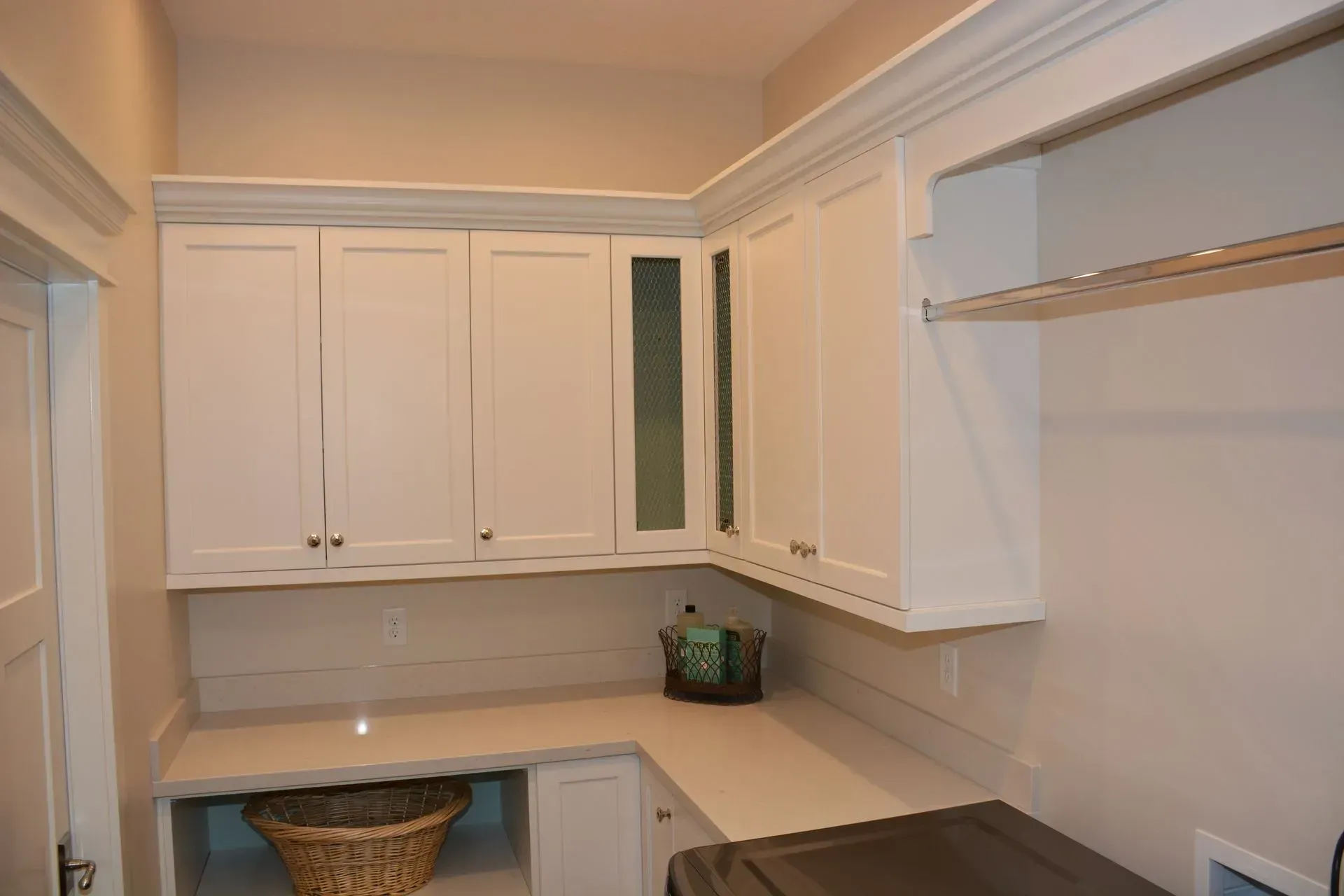 White kitchen cabinets installed above a countertop with a laundry basket below and a rack on the side wall.