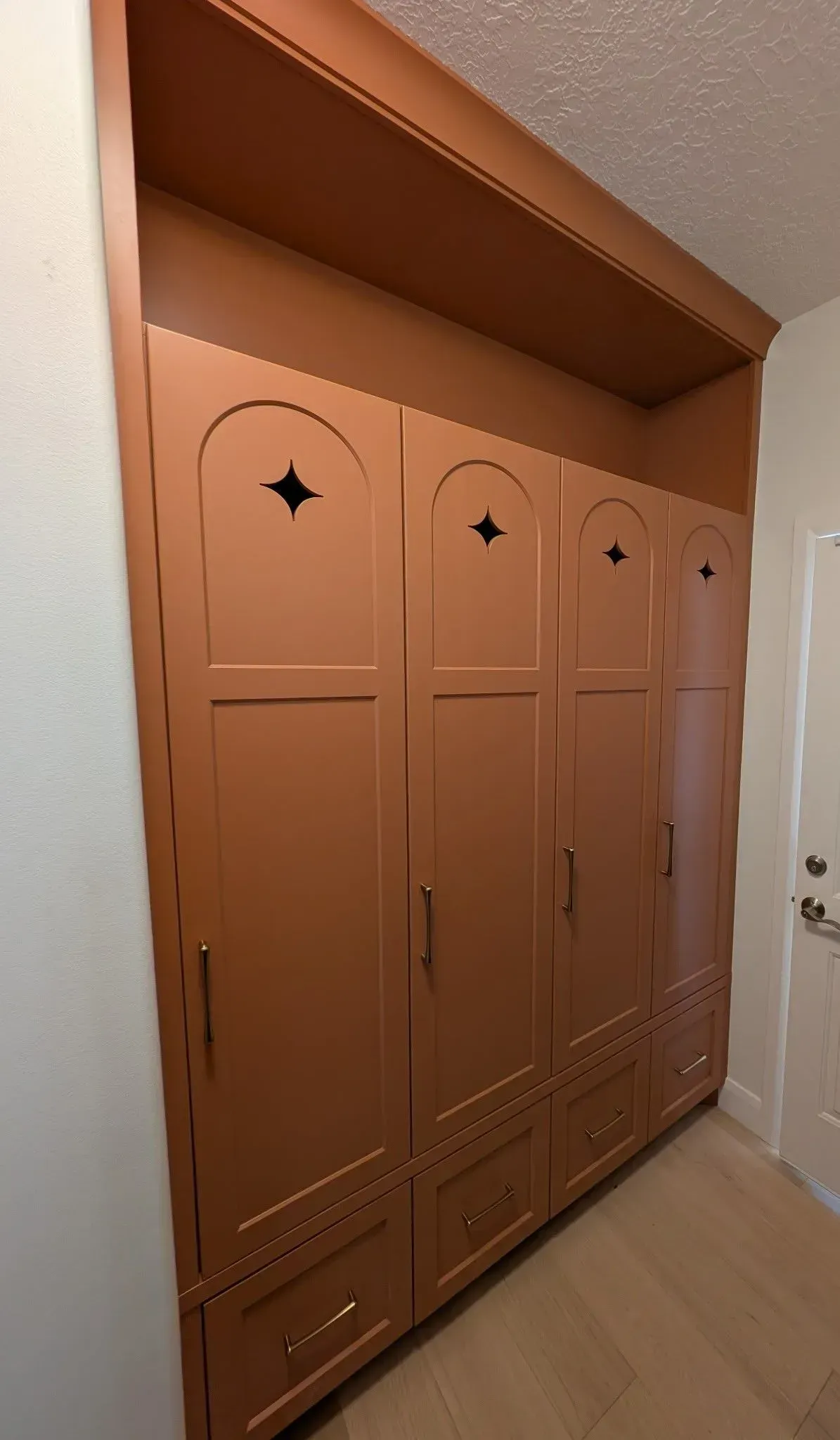 A wall of tall, terracotta-colored cabinets with arched details, diamond cutouts, and bottom drawers in a hallway.