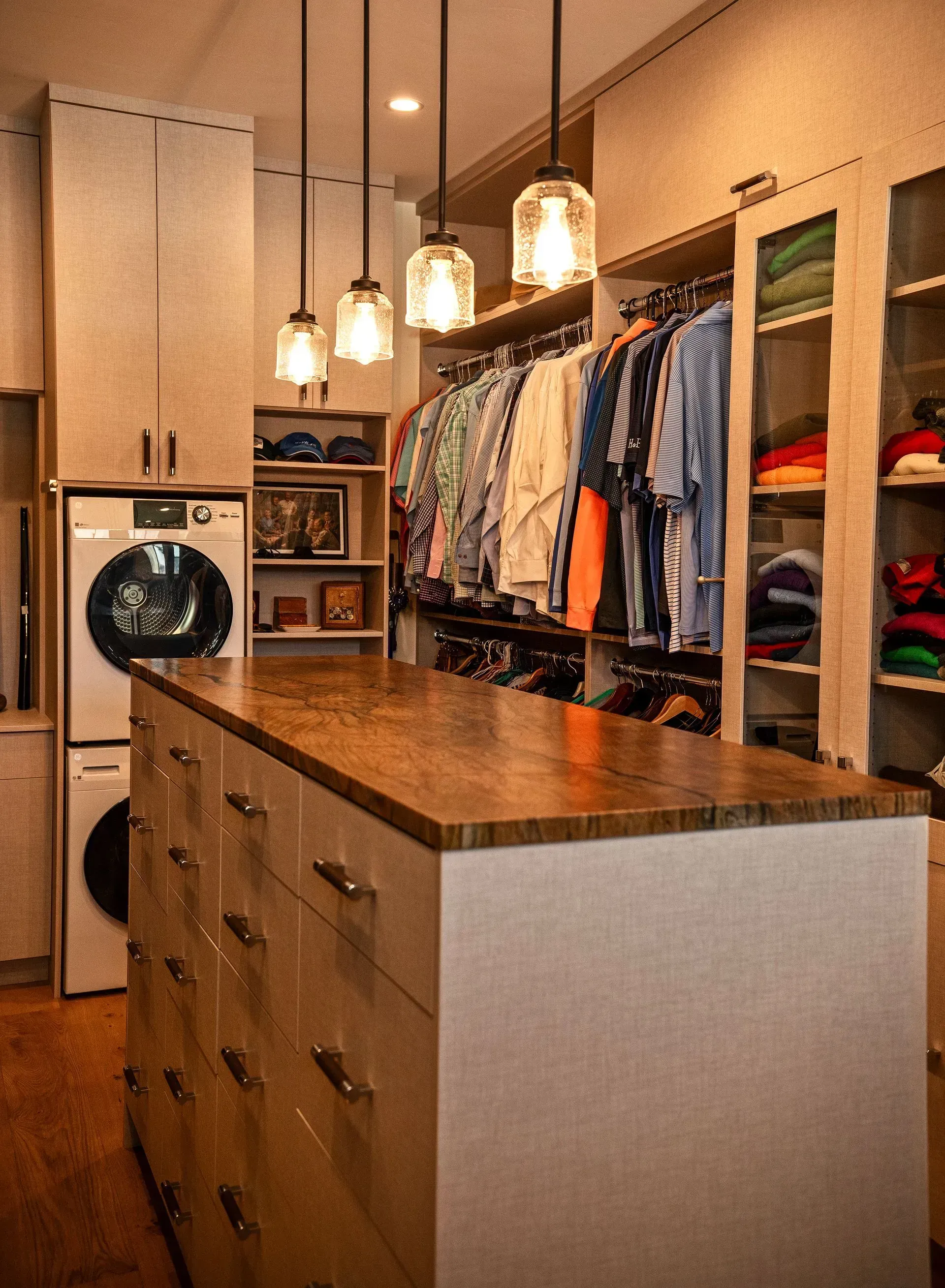 A walk-in closet with a laundry station, a central island with wood countertop and drawers, and pendant lights overhead.