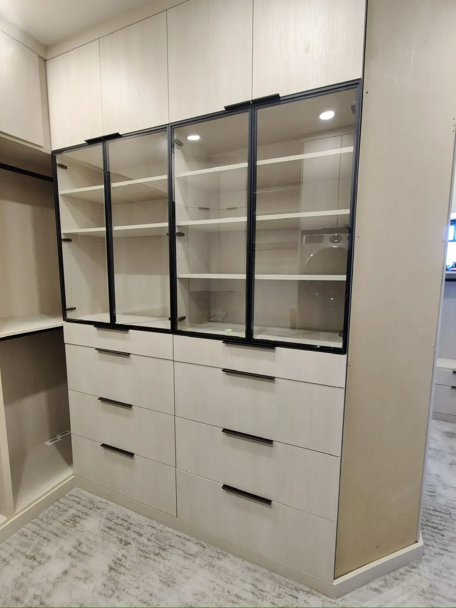 A custom closet featuring light wood cabinets with six drawers and glass-front shelving with black metal frames.