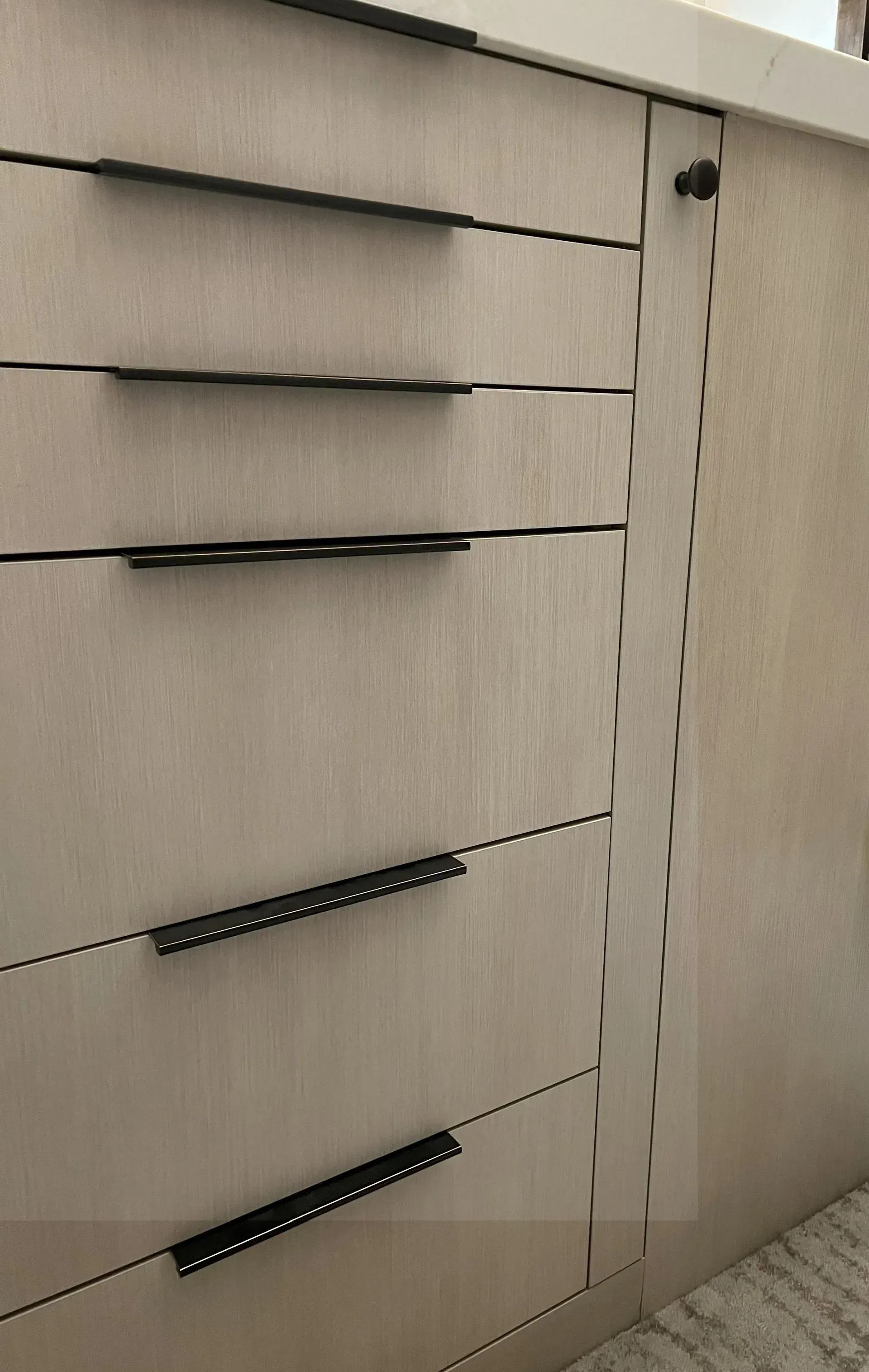 A close-up of a light-wood cabinet with five black, minimalist drawer pulls and a small, circular black knob on a door.