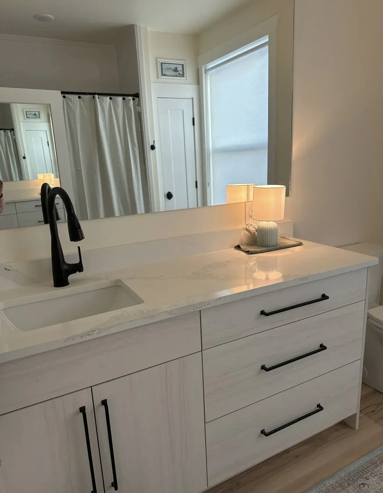 A modern bathroom vanity with a white marbled countertop, black faucet, and light wood cabinets, reflected in a mirror.