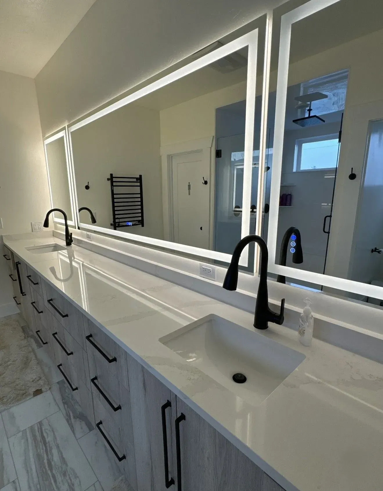 A modern bathroom vanity featuring a light wood finish, white countertop, two black faucets, and large, backlit mirrors.