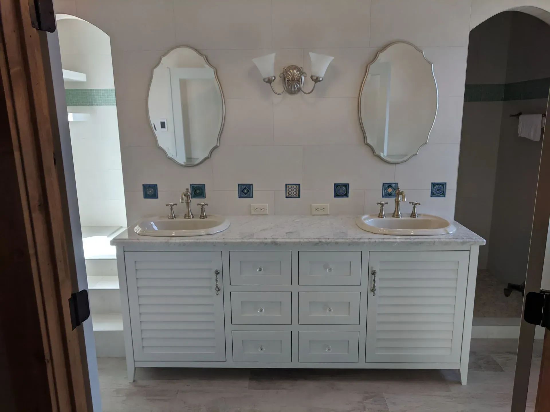 A double vanity with white cabinets, marble countertop, two oval mirrors, and light blue square tile accents.