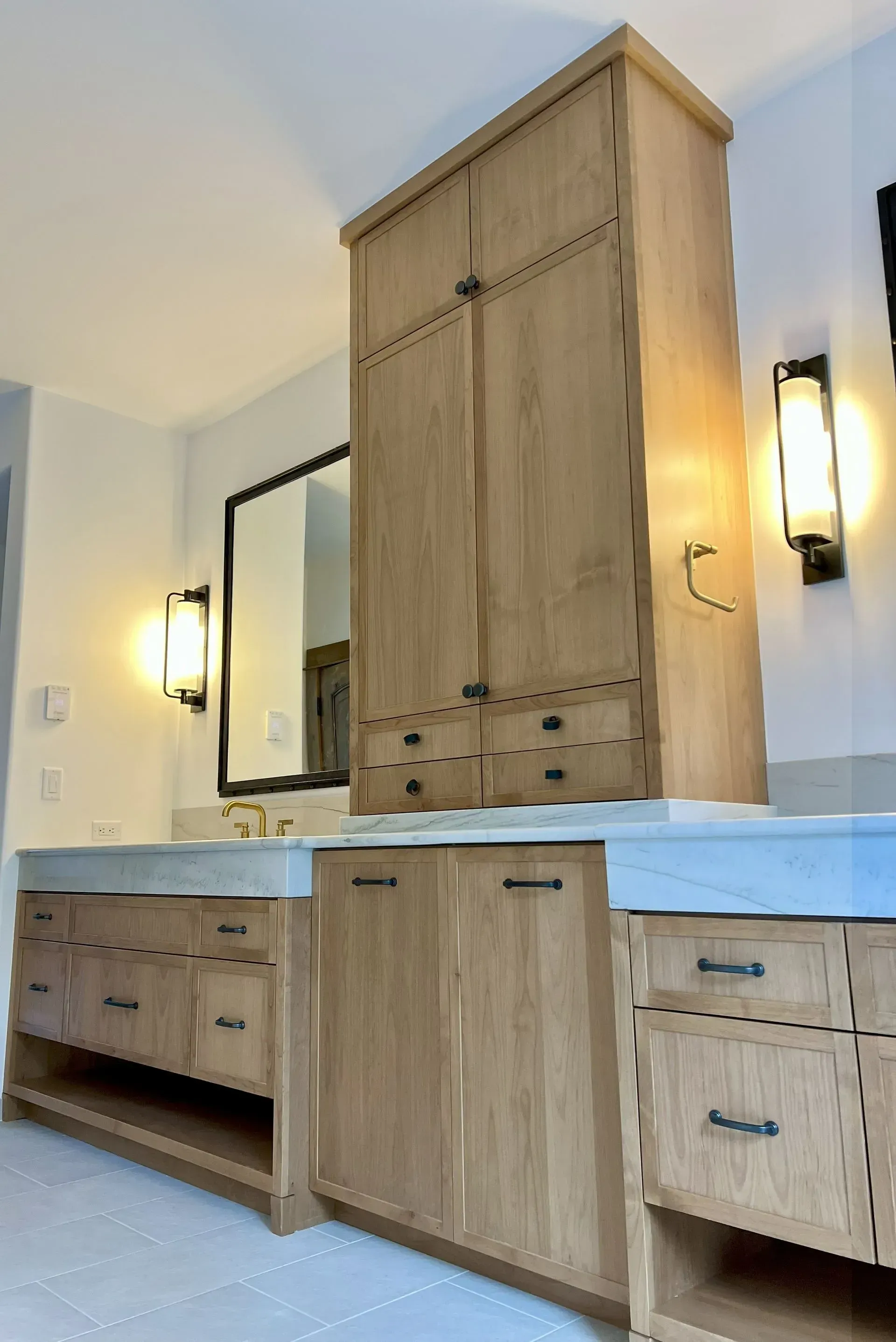 A bathroom vanity featuring light oak wood cabinets, a marble countertop, and wall-mounted lights on a white wall.