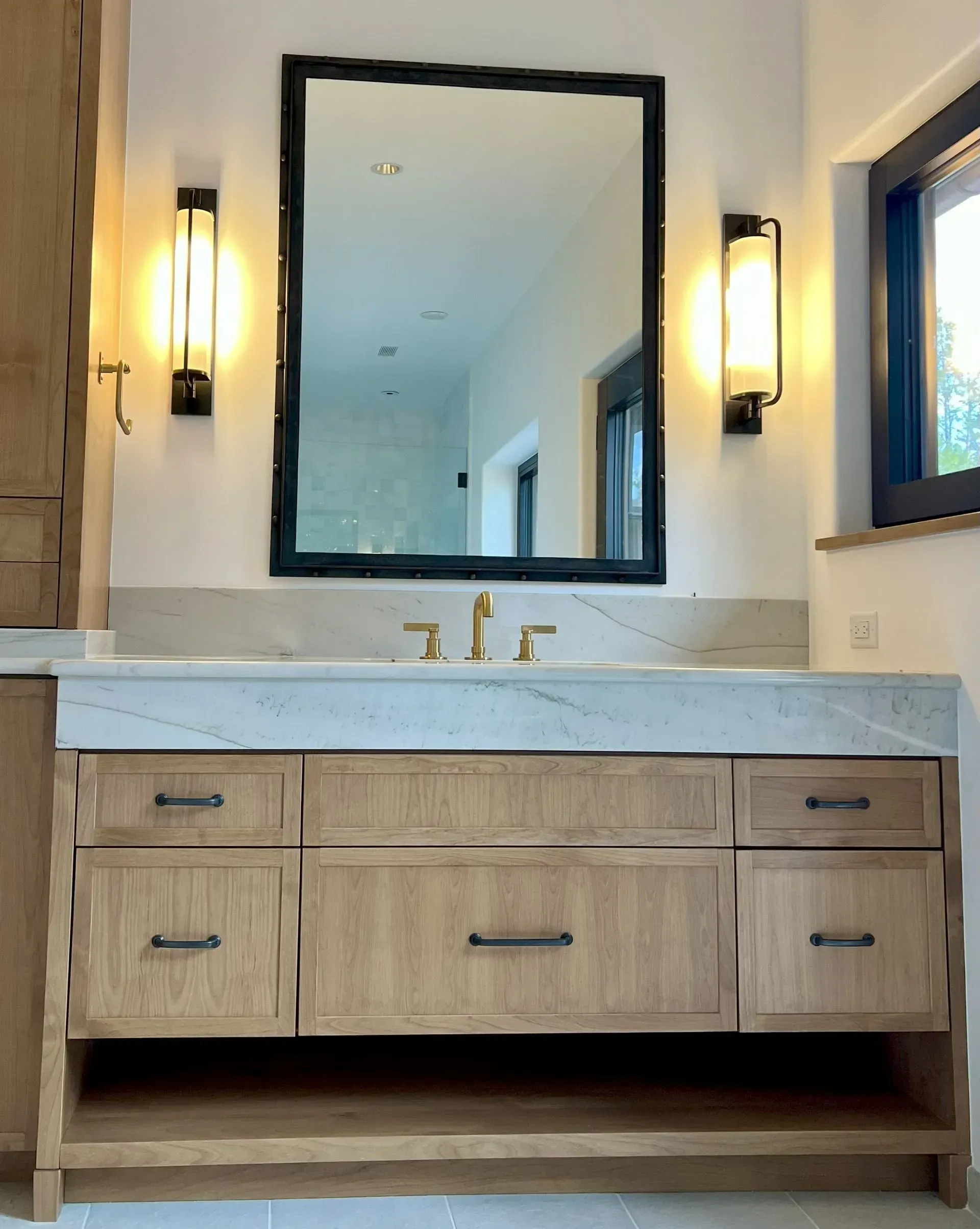 A light wood bathroom vanity with a white marble countertop, black-framed mirror, and vertical wall sconces.