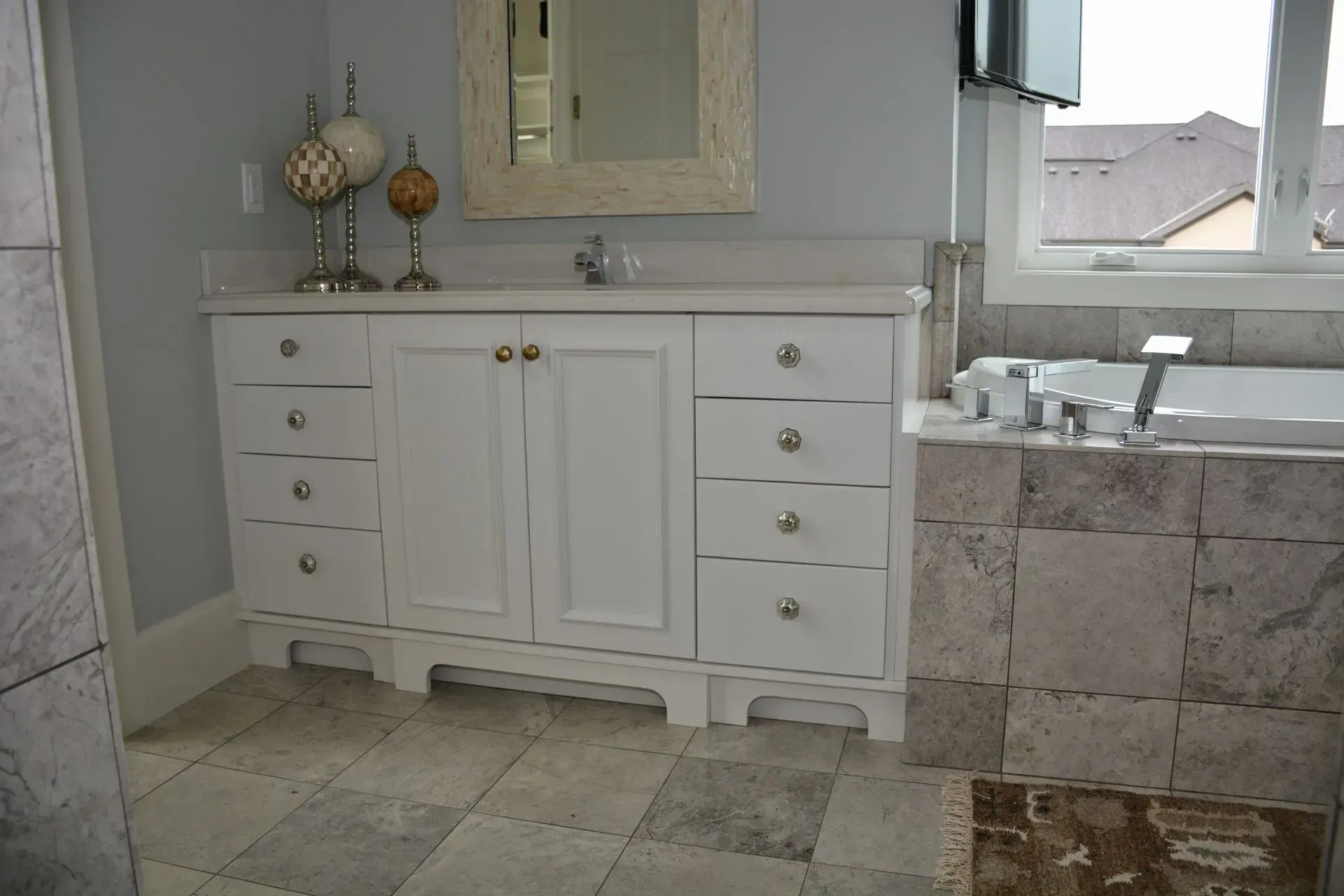 A white vanity with drawers and cabinet doors next to a tiled bathtub in a bathroom with light gray walls.