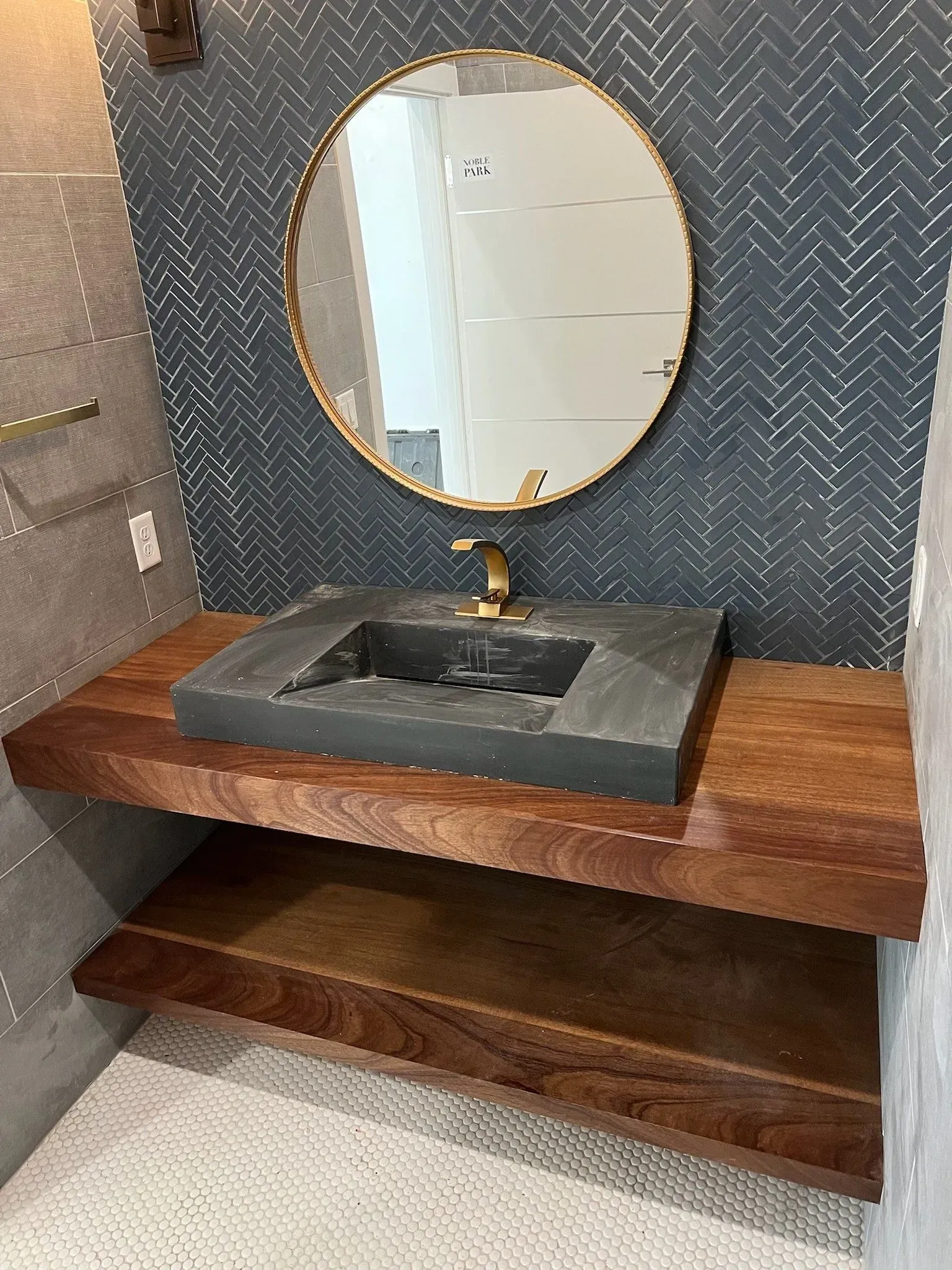 A bathroom vanity featuring a dark stone sink, wooden shelves, a gold faucet, and a round mirror on herringbone wallpaper.