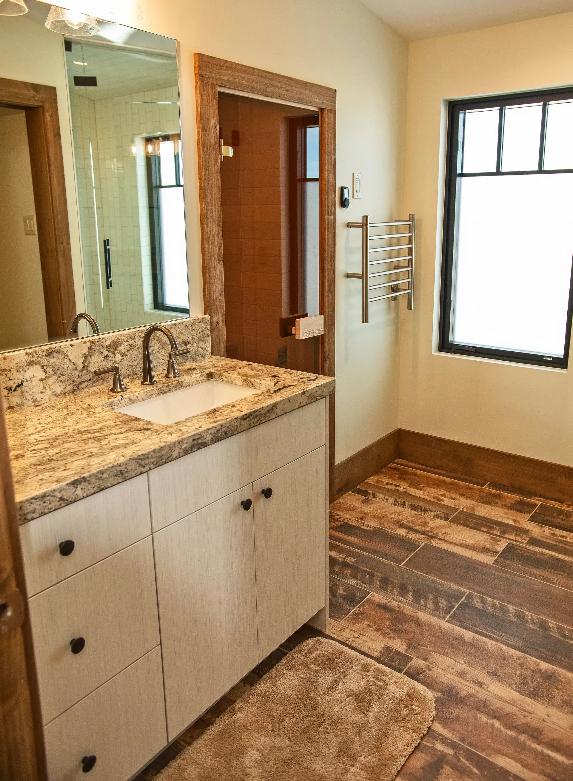 A rustic bathroom with a speckled granite countertop, light wood vanity, wall mirror, towel rack, and textured floor.