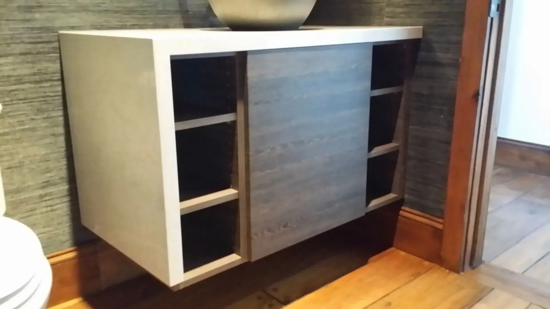 A wall-mounted, white-topped bathroom vanity featuring open side shelving and a grey wood-grain sliding center cabinet.