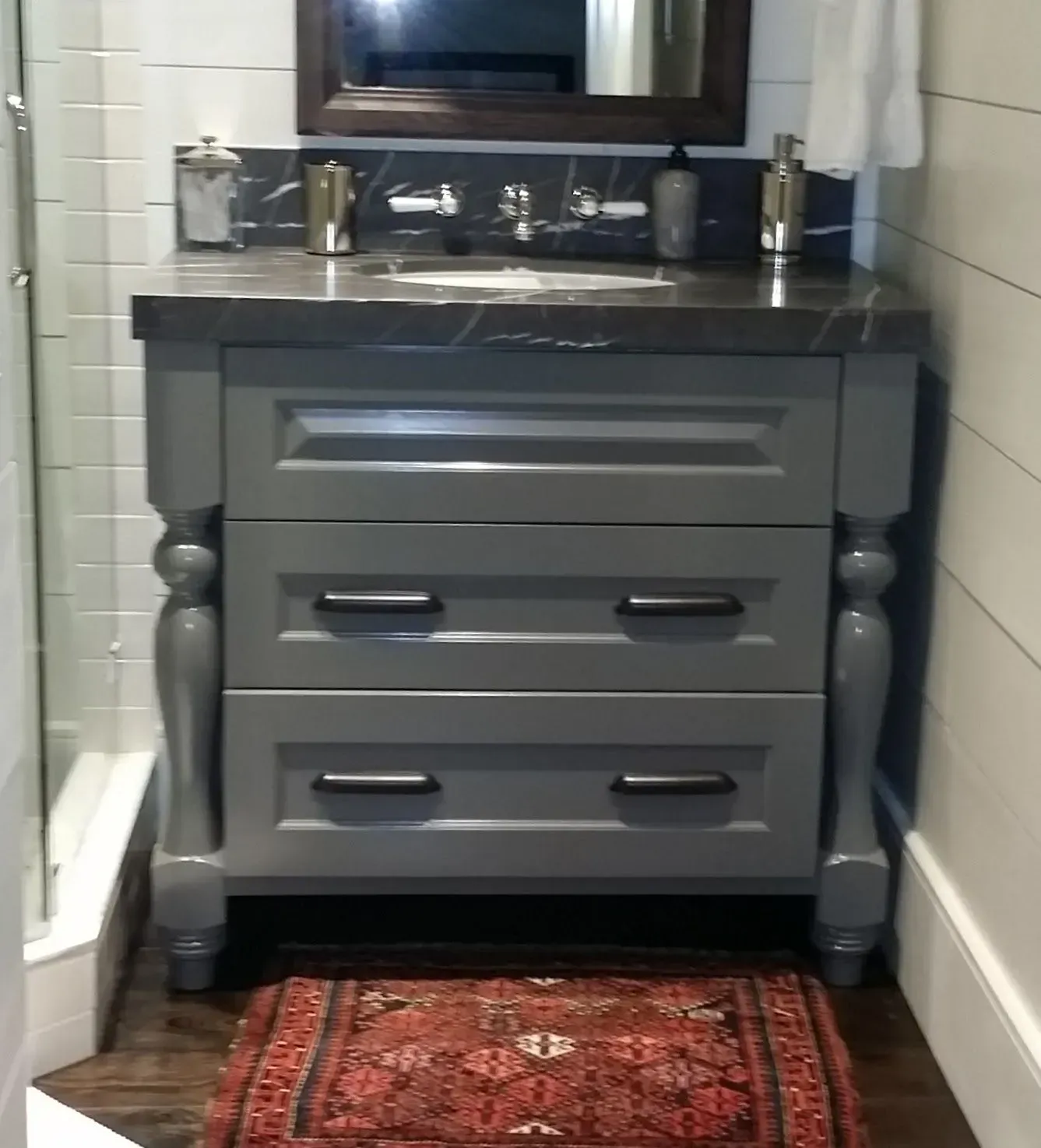 A grey bathroom vanity with a dark marble top, black hardware, and a small red patterned rug on the floor.