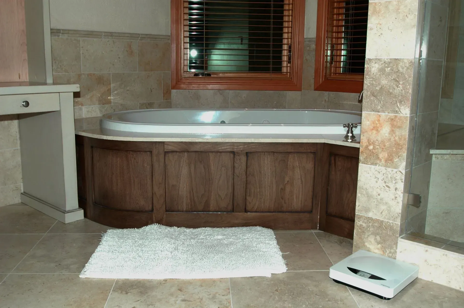 A bathroom featuring a jetted tub with a wood-paneled surround, a white bath mat, and a scale on a tiled floor.