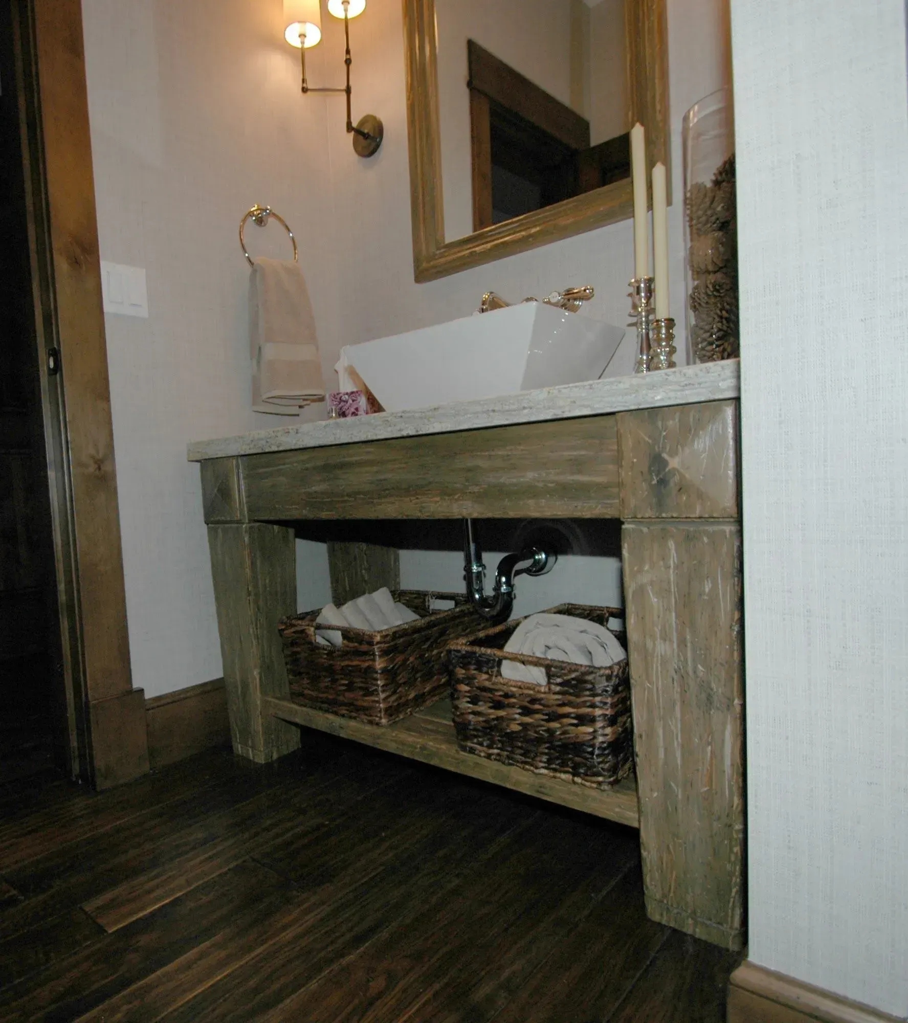 A rustic wood vanity with a stone countertop, white vessel sink, and two storage baskets underneath in a small bathroom.