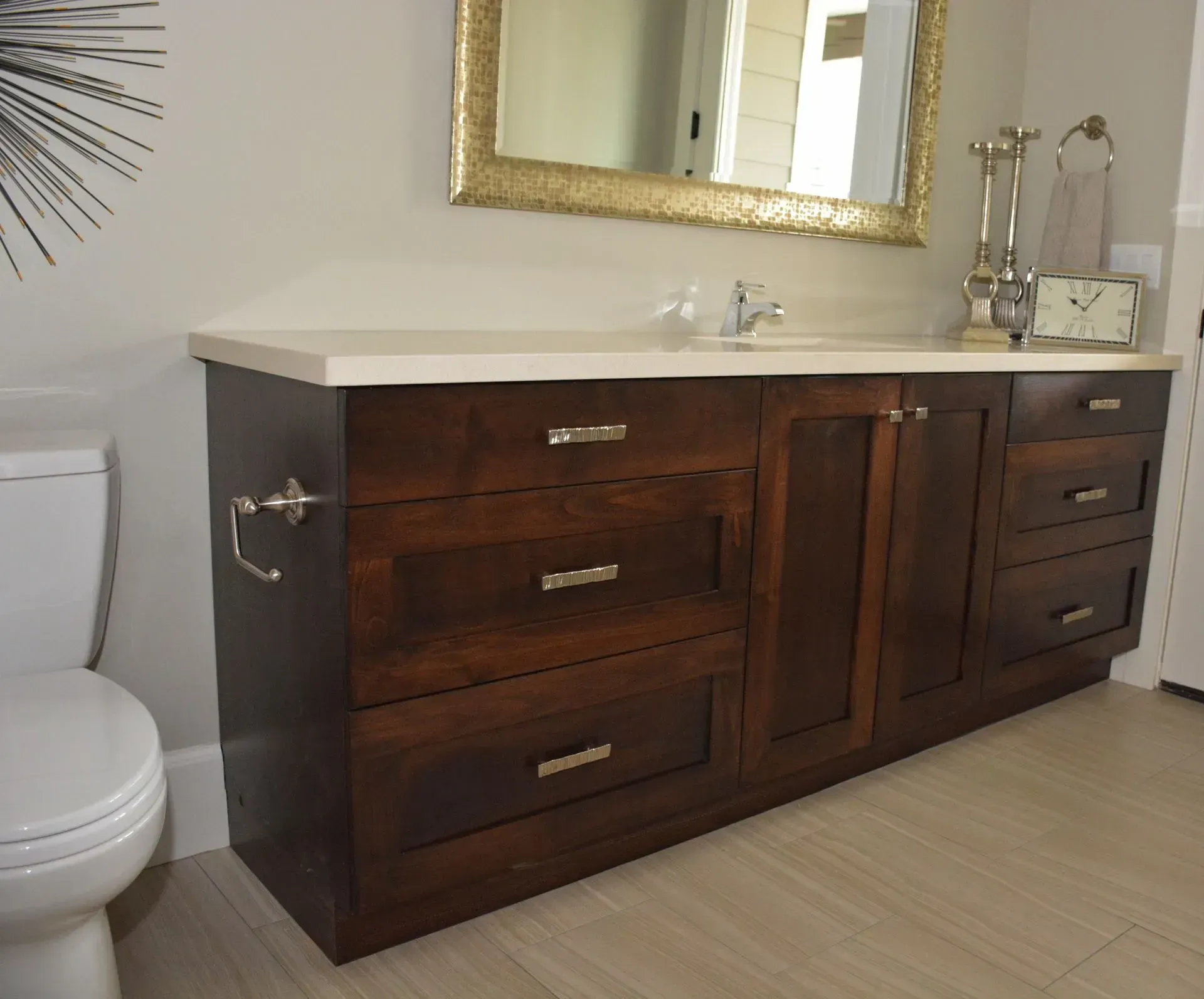 A dark wood bathroom vanity with a light-colored countertop and a gold-framed mirror sits next to a white toilet.