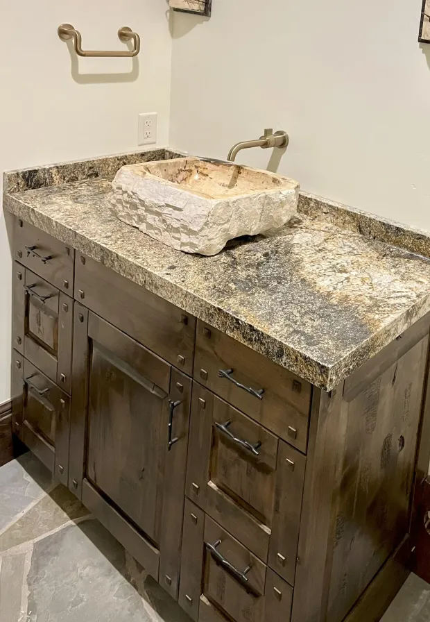A rustic bathroom vanity with a granite countertop, stone vessel sink, and brass faucet against a cream wall.