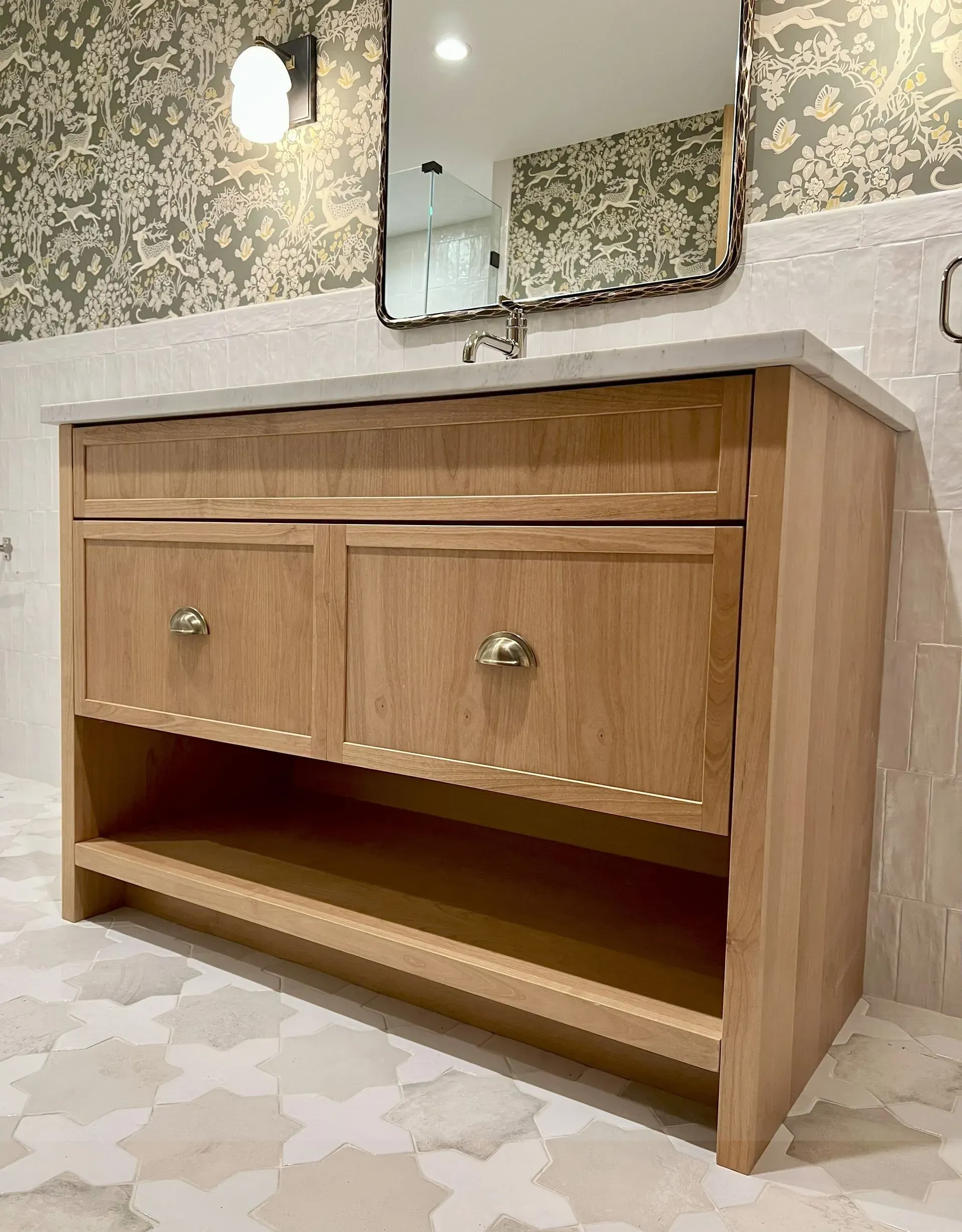 A wooden bathroom vanity with two cabinets and an open bottom shelf, situated below a wall mirror and patterned wallpaper.