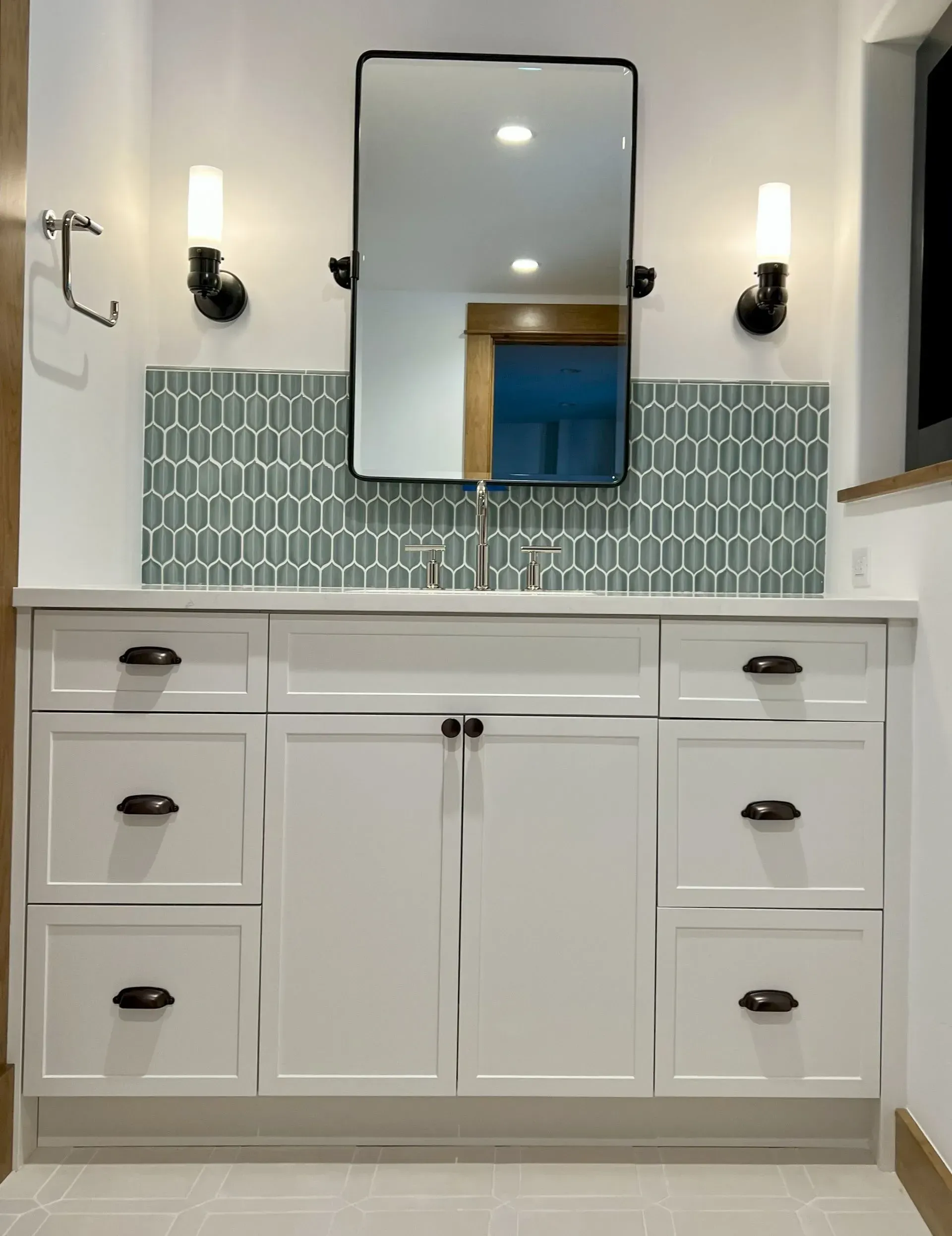 A white bathroom vanity with dark hardware, a rectangular mirror, two wall sconces, and a blue-green patterned backsplash.