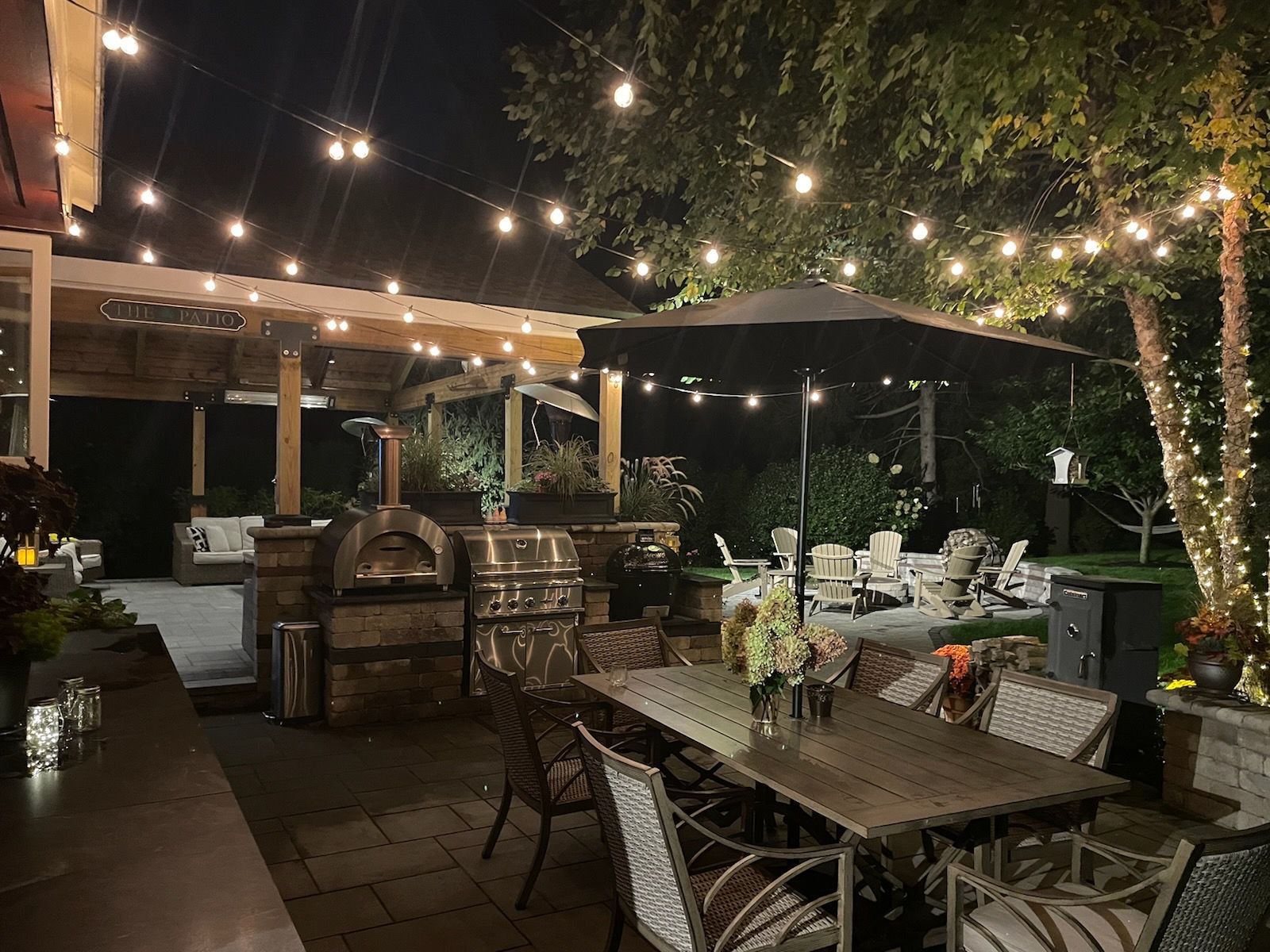 A patio with a table , chairs , umbrellas and a pizza oven at night.