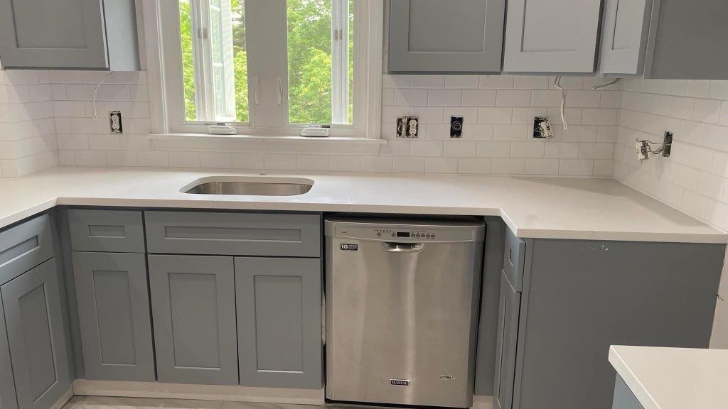 A kitchen with gray cabinets , white counter tops , a sink and a dishwasher.