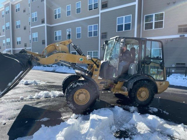 A cat wheel loader is clearing snow from a parking lot in front of a building.