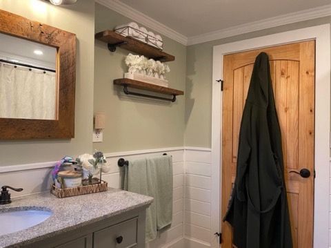 A bathroom with a sink , mirror , shelves and a robe hanging on the door.