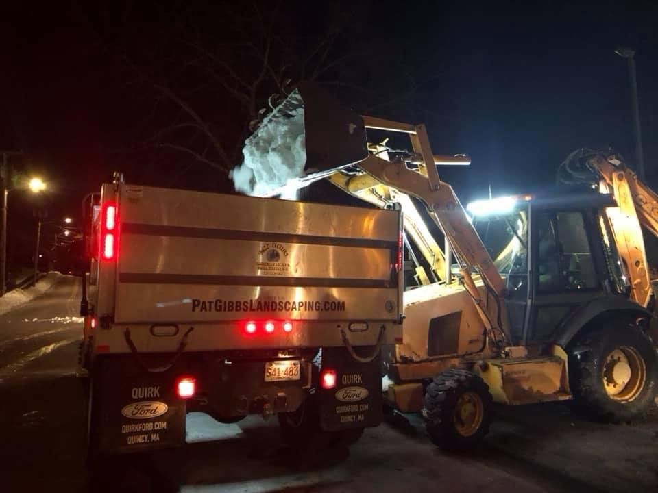 A dump truck is being loaded with snow by a bulldozer at night.