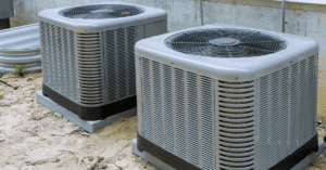 Two gray air conditioning units on a sandy surface.