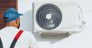 Man in overalls and blue cap inspecting an outdoor air conditioning unit mounted on a white wall.