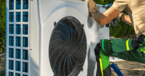 Technician installing an outdoor heat pump unit; using a drill, wearing gloves and green work pants.