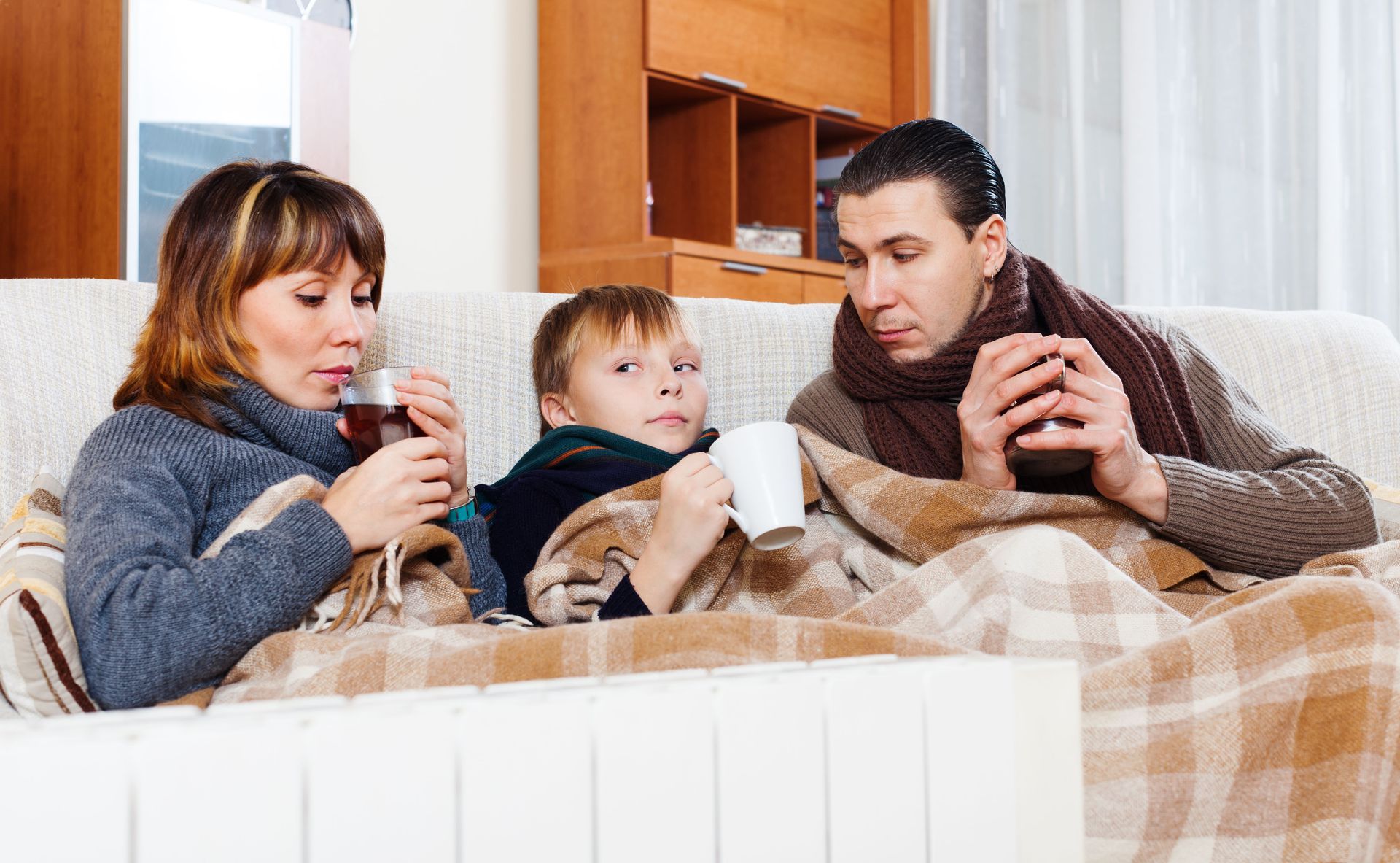 Family wrapped in blankets on a couch, drinking from mugs; looks like they have a cold.