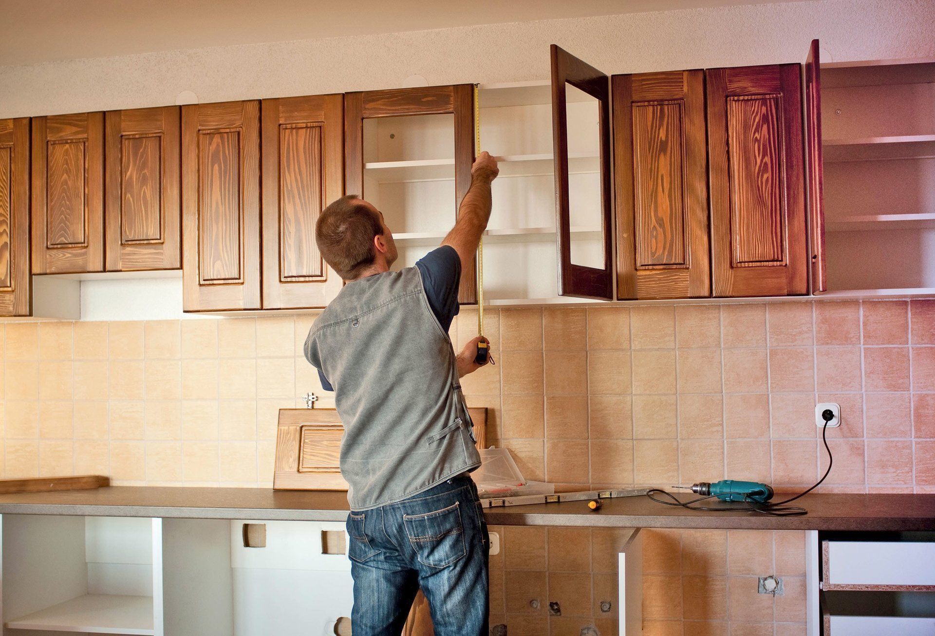 A man is working on a kitchen cabinet with a drill.