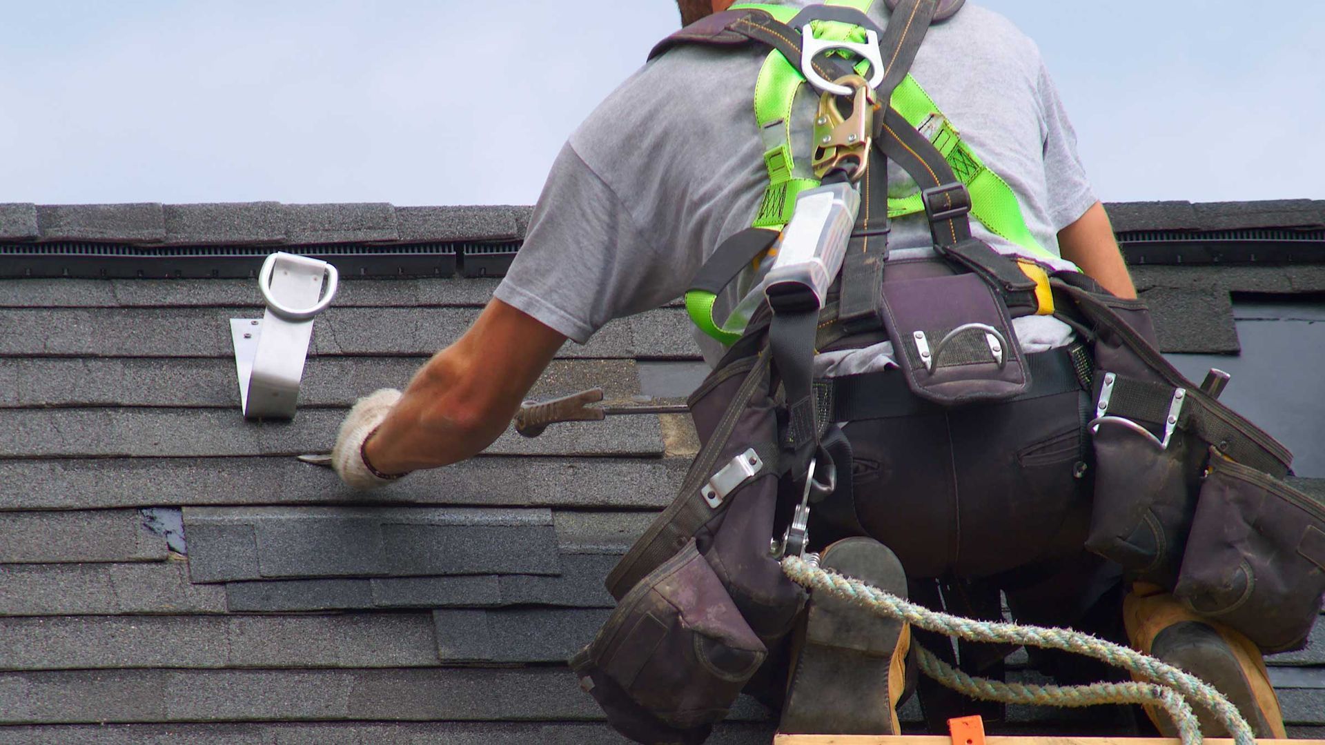 Roofer on a roof installing shingles, wearing safety harness.
