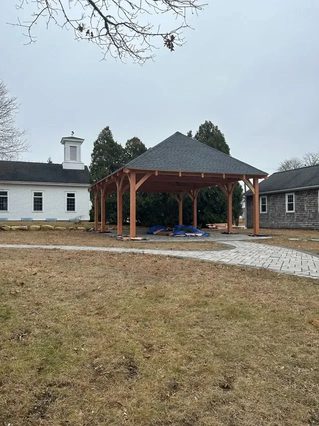 Wooden gazebo with dark roof, in a grassy yard near white buildings. Overcast day.
