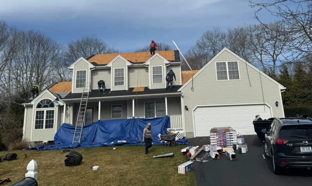 Roofers working on a house roof, supplies on the ground. Blue tarp, sunny day.
