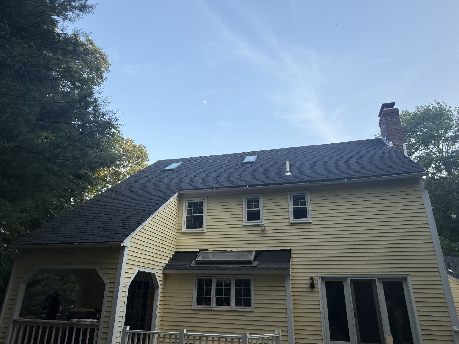Yellow house with dark roof, windows, and chimney against a blue sky, surrounded by trees.