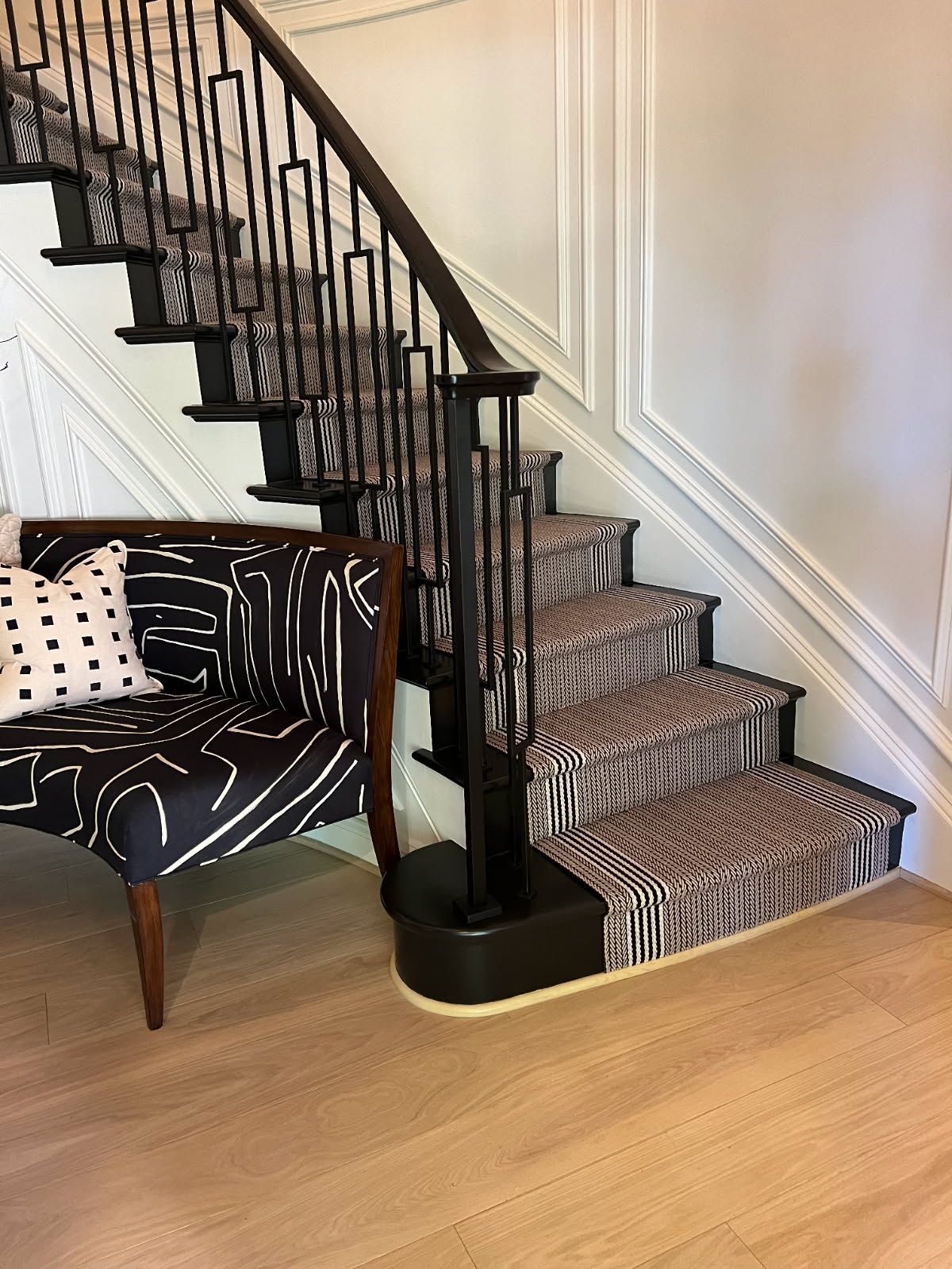 Staircase with black iron railing, patterned carpet, and a black and white patterned chair. Light wood floor.