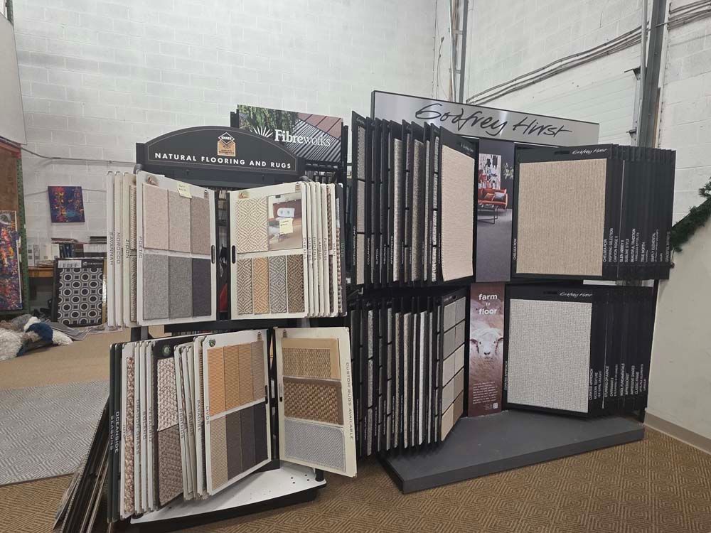 Display of various carpet samples in a showroom, with a rug and artwork visible in the background.