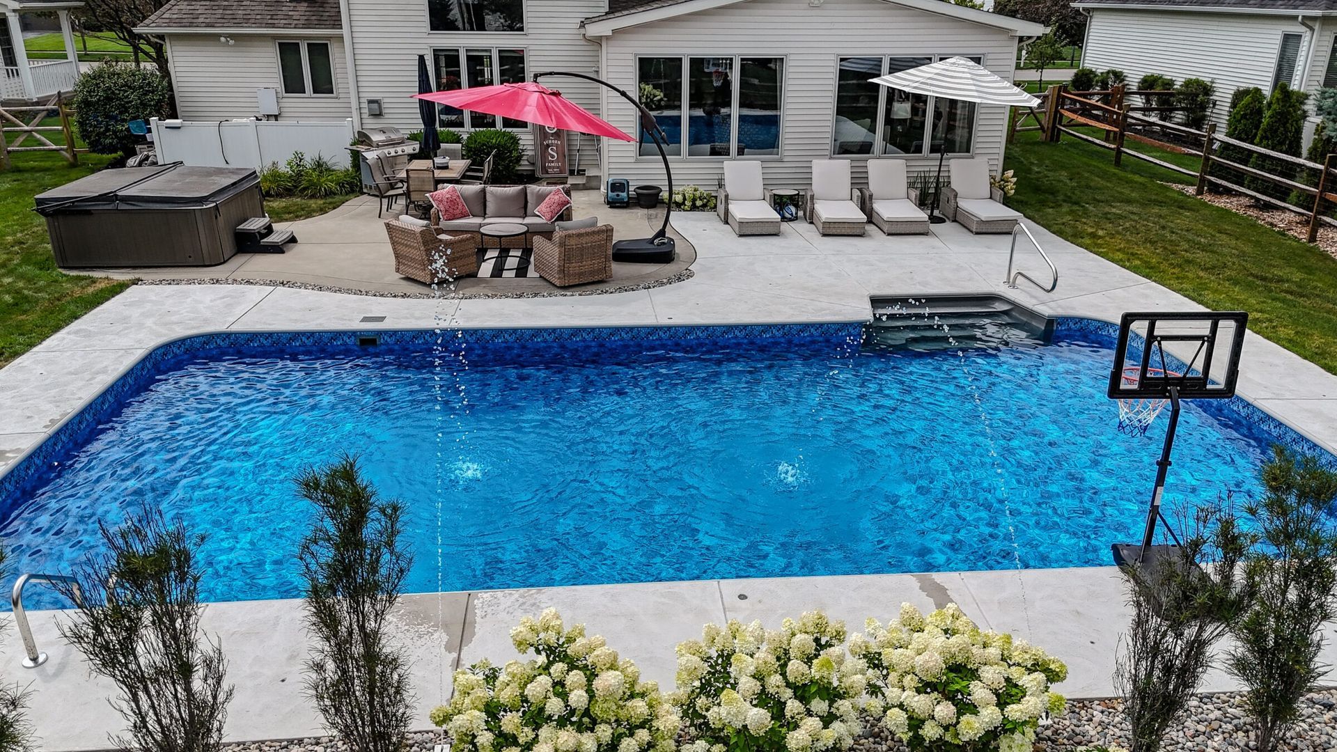Rectangular vinyl liner pool with vibrant blue water and built-in deck jets, surrounded by a concrete patio. The setup includes lounge chairs under striped umbrellas, a cozy seating area with a pink umbrella, a hot tub, and a basketball hoop at one end. Lush landscaping and flowering shrubs add a welcoming touch to the backyard space.