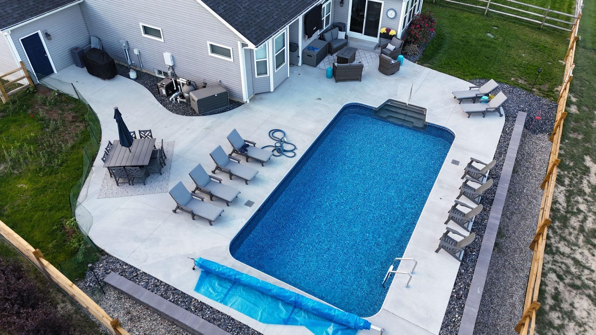 An overhead view of a rectangular pool with blue water, surrounded by a spacious concrete deck. Multiple lounge chairs and a dining set with an umbrella are arranged neatly around the pool.