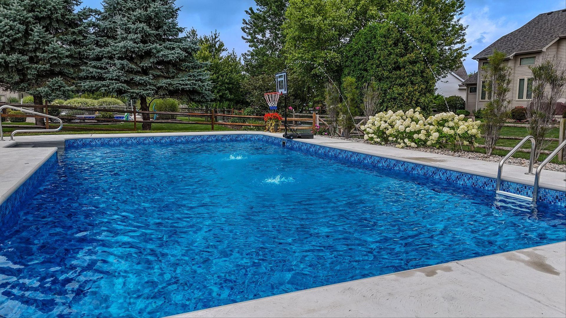 A rectangular swimming pool with fountains spraying into the water, bordered by concrete decking. A hot tub, lounge chairs, and wicker patio furniture sit nearby, with a large white house in the background.