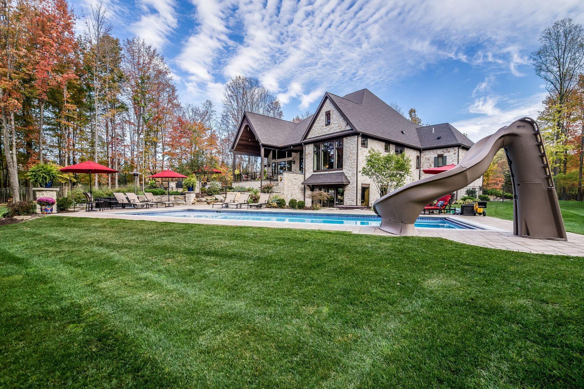 Elegant backyard scene with a rectangular inground pool, large waterslide, and professionally manicured lawn, set against a stone-clad luxury home and vibrant fall foliage.