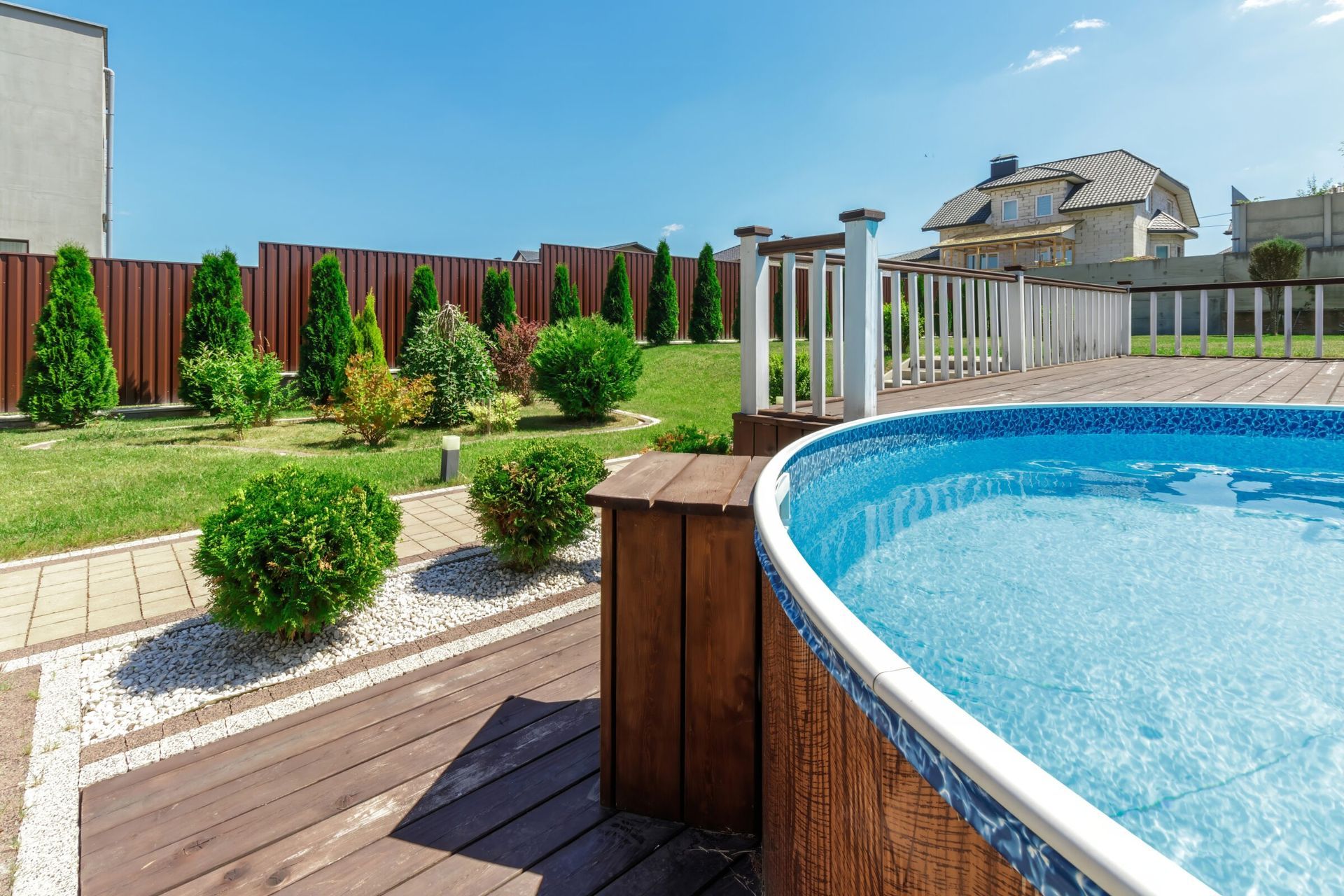 Above-ground pool with a wooden deck and white railing in a neatly landscaped backyard featuring paved walkways, decorative gravel, and small evergreen shrubs, all enclosed by a tall red privacy fence and backed by a large suburban house.
