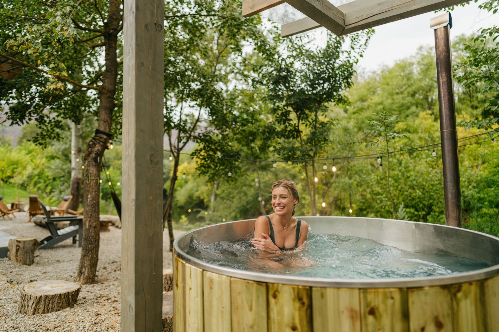 Smiling woman relaxes in a round wooden stock tank-style pool surrounded by trees, string lights, and rustic outdoor seating on a gravel patio in a natural, wooded setting.